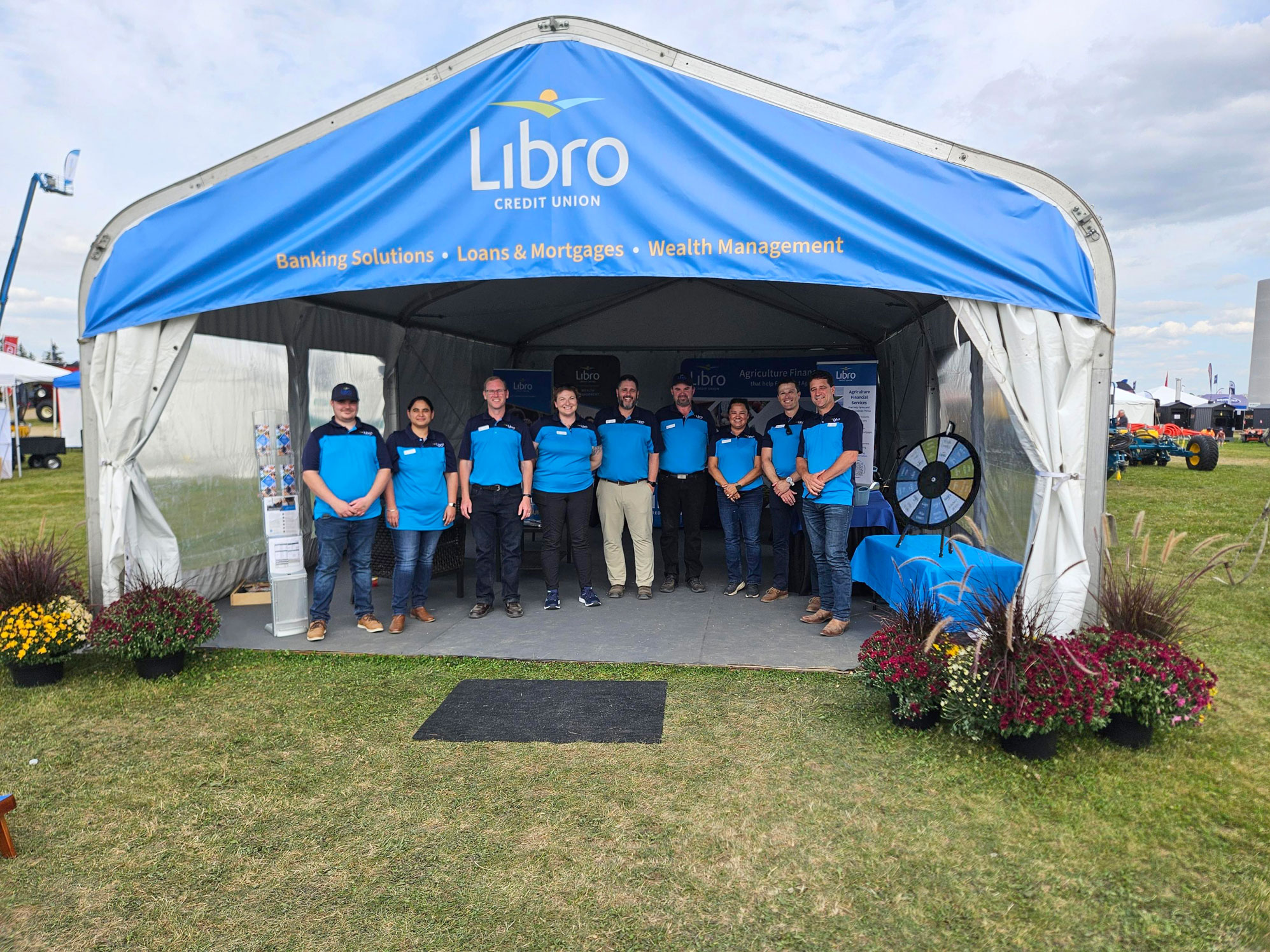Nine people wearing blue shirts stand in front of a Libro Credit Union tent at an outdoor event, surrounded by potted flowers and informational displays.