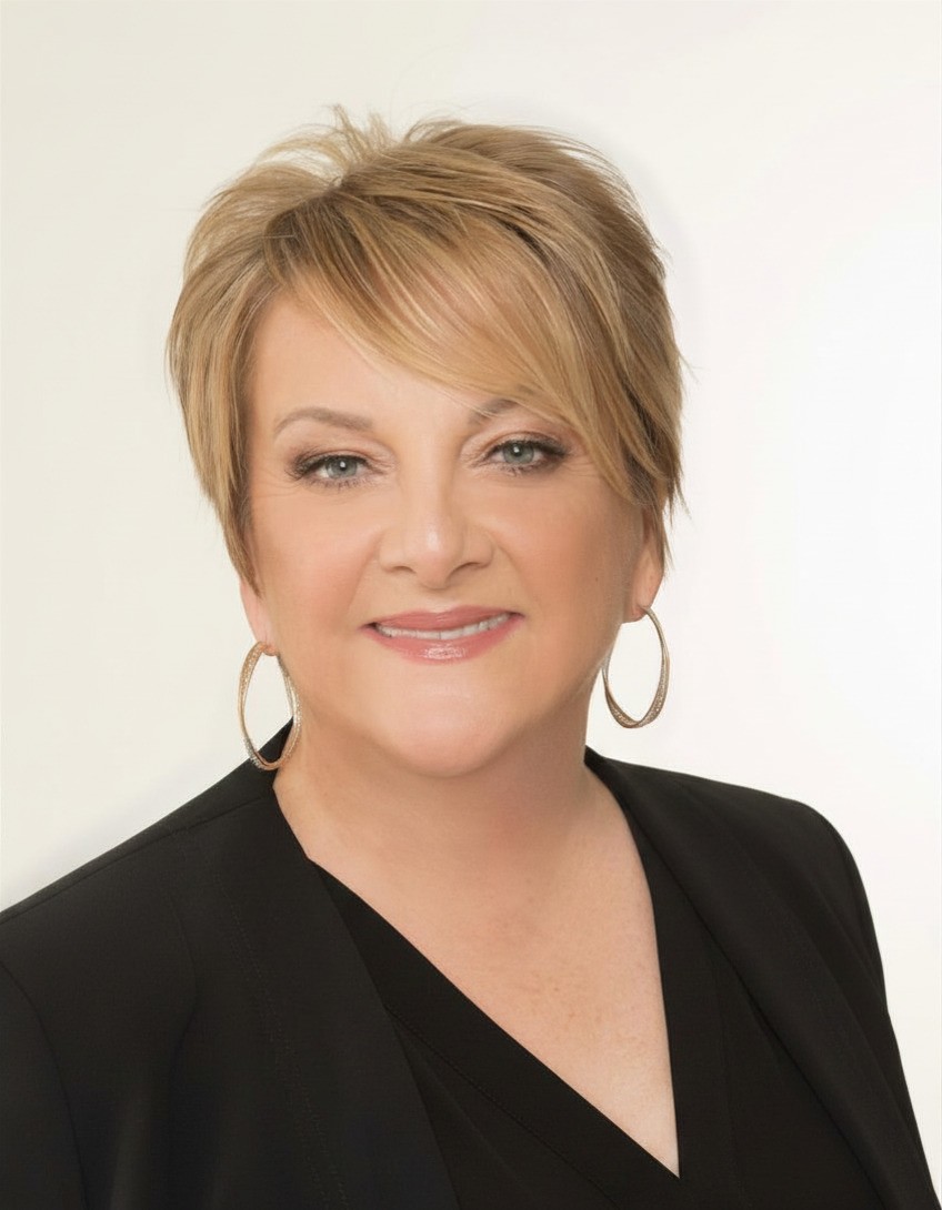 A woman with short, light brown hair wearing hoop earrings and a black blazer smiles at the camera against a plain background.