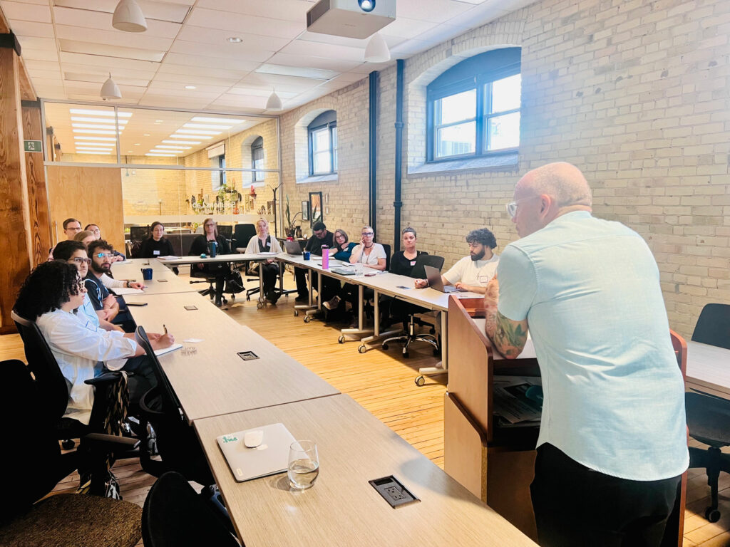A bald man in a light blue shirt speaks at a podium to a group of people seated around tables in a bright, modern meeting room with brick walls.