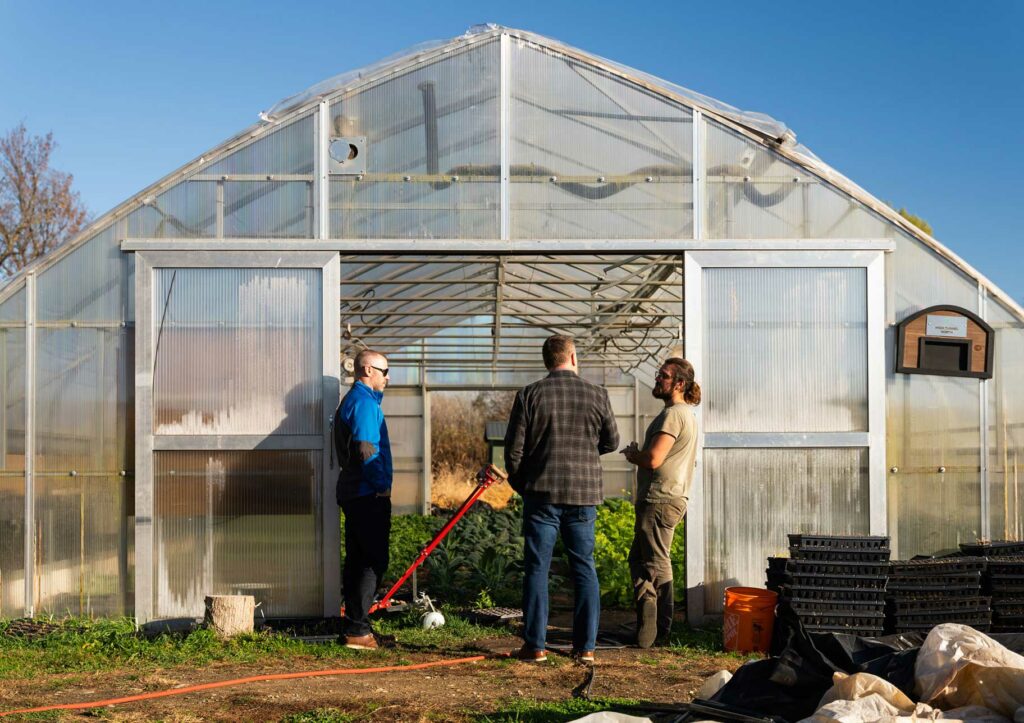 Three people stand and talk outside the entrance of a greenhouse on a sunny day, with plants visible inside and gardening supplies nearby.