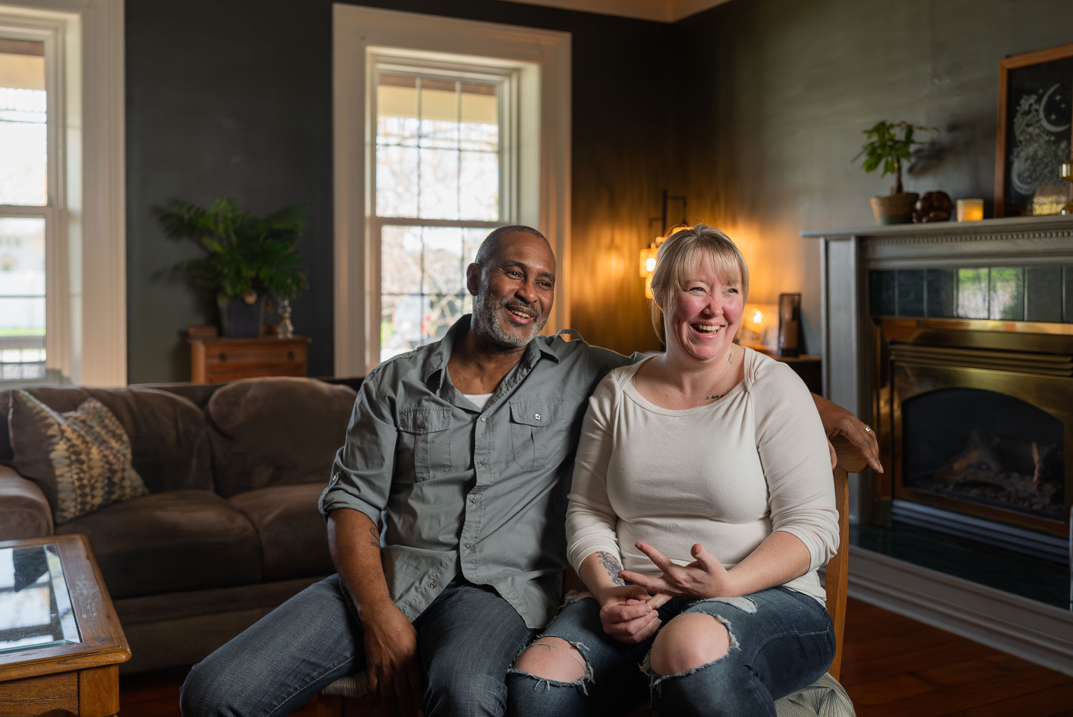 A man and woman sit together on a chair in a cozy living room, both smiling. The room features large windows, a couch, and a fireplace with decorative items.