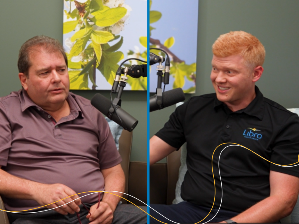 Two men sit in a studio setting with microphones; one wears a purple shirt, the other a black Libro shirt. A leafy artwork hangs on the wall behind them.