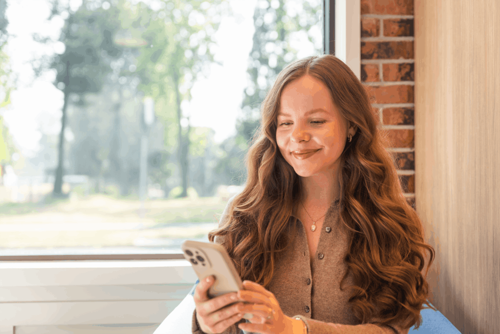 A woman with long, wavy hair sits indoors near a window, smiling while looking at her smartphone.