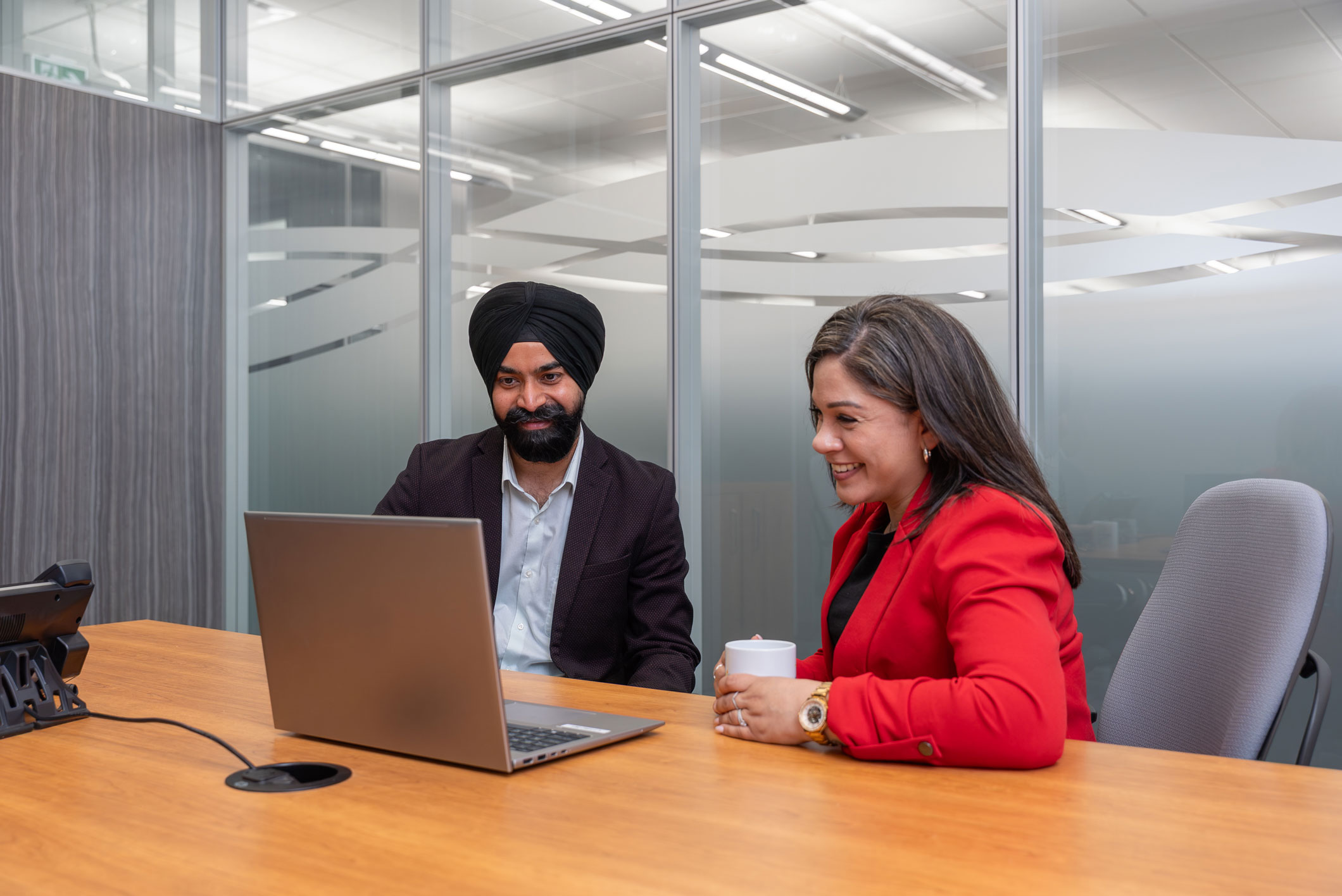 Two people sit at a conference table, looking at a laptop screen. The man wears a turban and suit; the woman wears a red blazer and holds a white mug.
