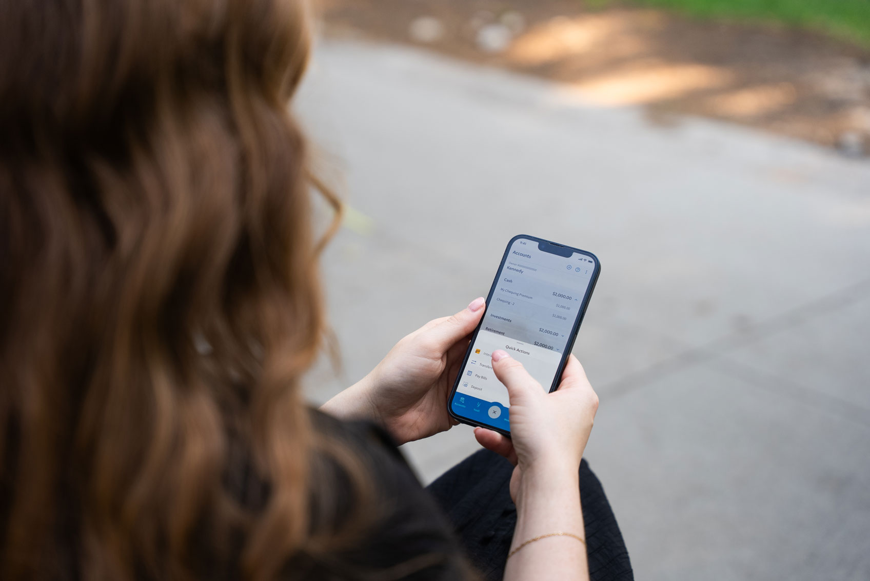 Person with long hair using a smartphone outdoors, scrolling through a financial or banking app on the screen.