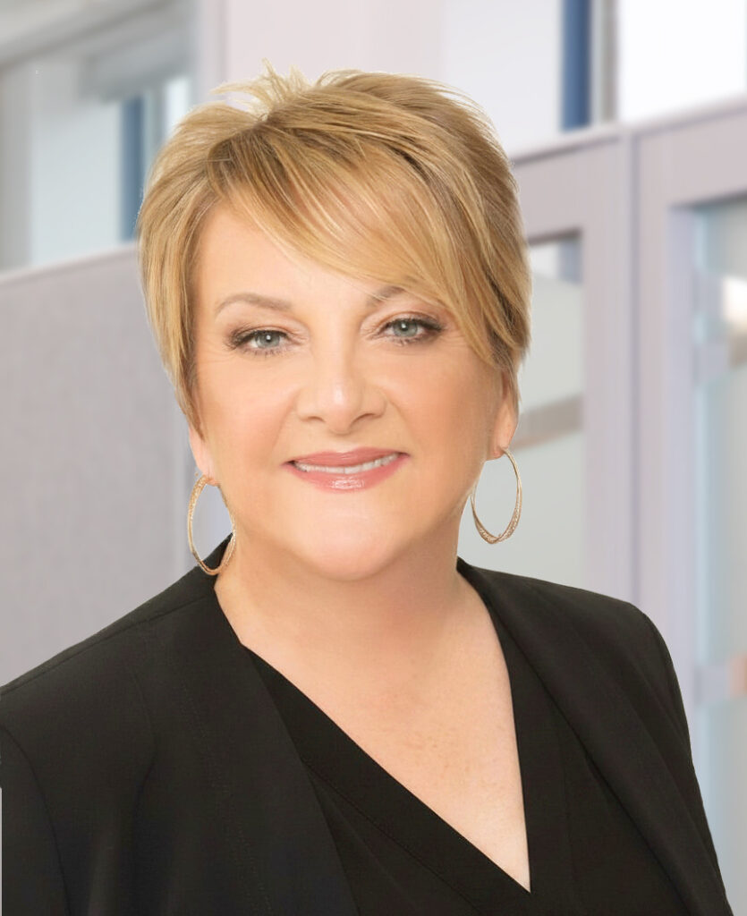 Woman with short blonde hair, wearing hoop earrings and a black blazer, smiles at the camera in a modern office setting.