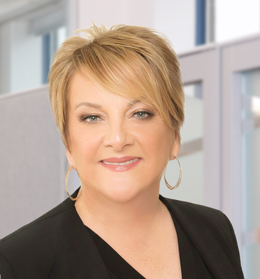 Woman with short blonde hair, wearing hoop earrings and a black blazer, smiles at the camera in a modern office setting.