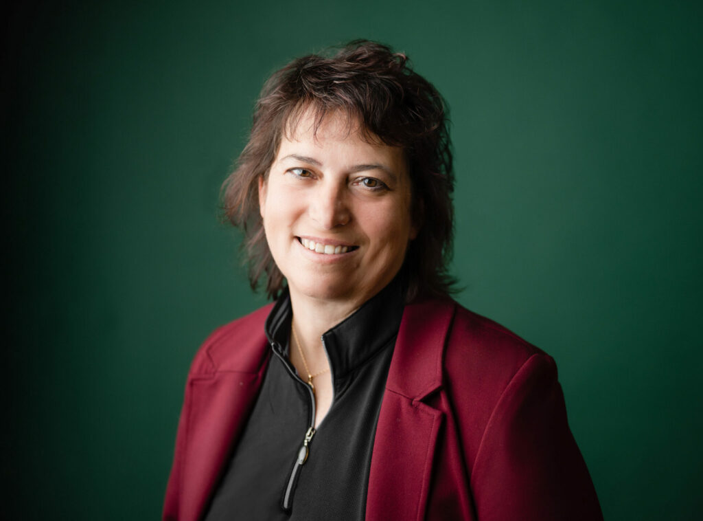 Smiling person with short brown hair, wearing a burgundy blazer over a black shirt, poses in front of a plain dark green background.