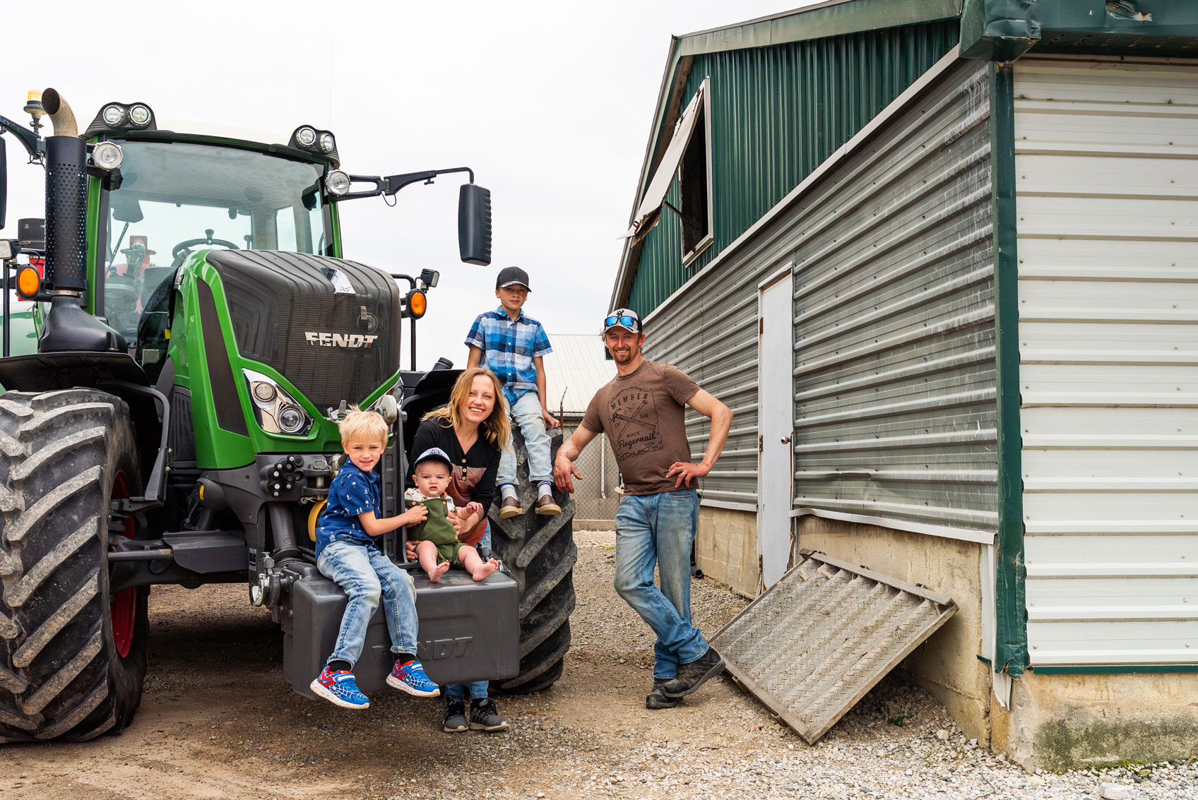 A family of five poses beside a green tractor and a metal barn on a farm. Two children sit on the tractor, while the others and two adults stand and smile near the barn.