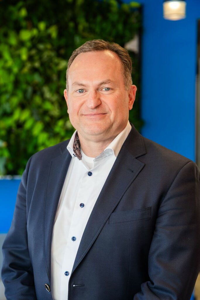 Man in a dark suit and white shirt stands in front of a blue wall and green leafy background, facing the camera with a neutral expression.