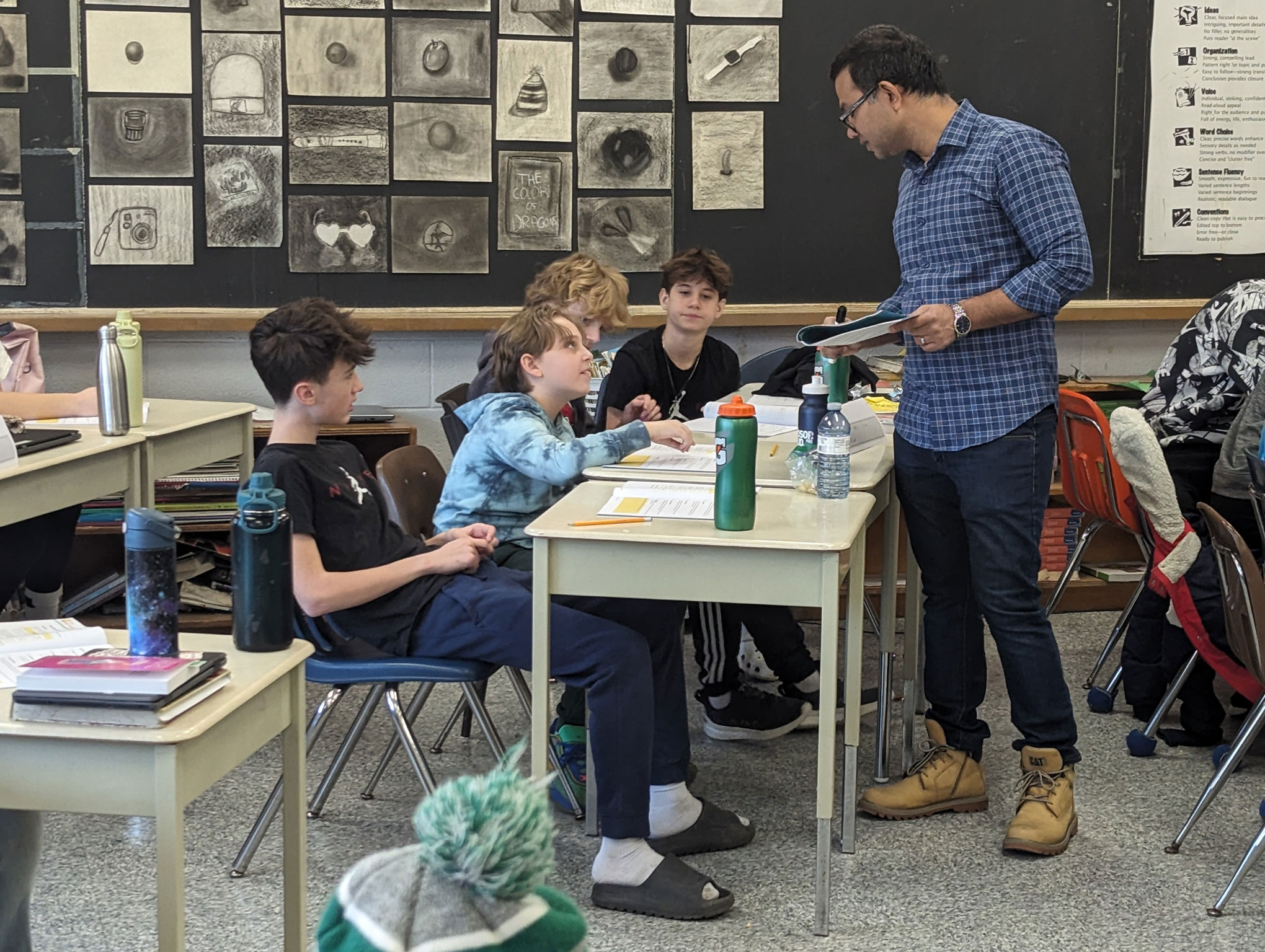 A teacher stands beside a table talking to three students working on assignments in a classroom with art displayed on the back wall.