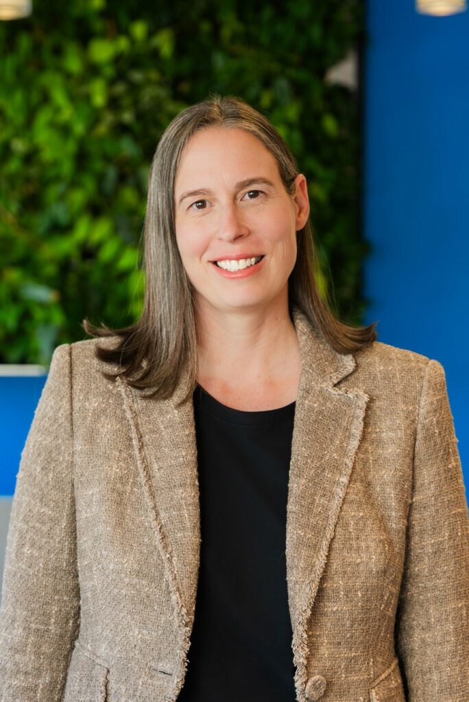 A woman with straight, shoulder-length brown hair, wearing a tan blazer and black shirt, stands smiling in front of a green plant wall and blue background.
