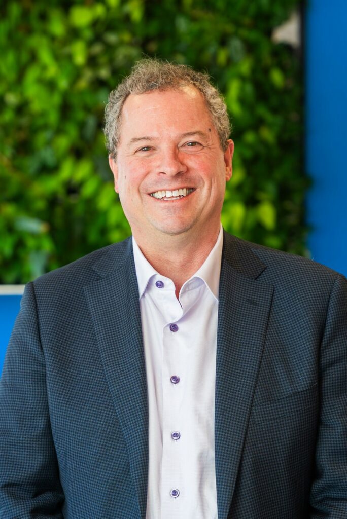 A man in a blue checked blazer and white shirt smiles at the camera, standing in front of a green leafy background and a blue wall.