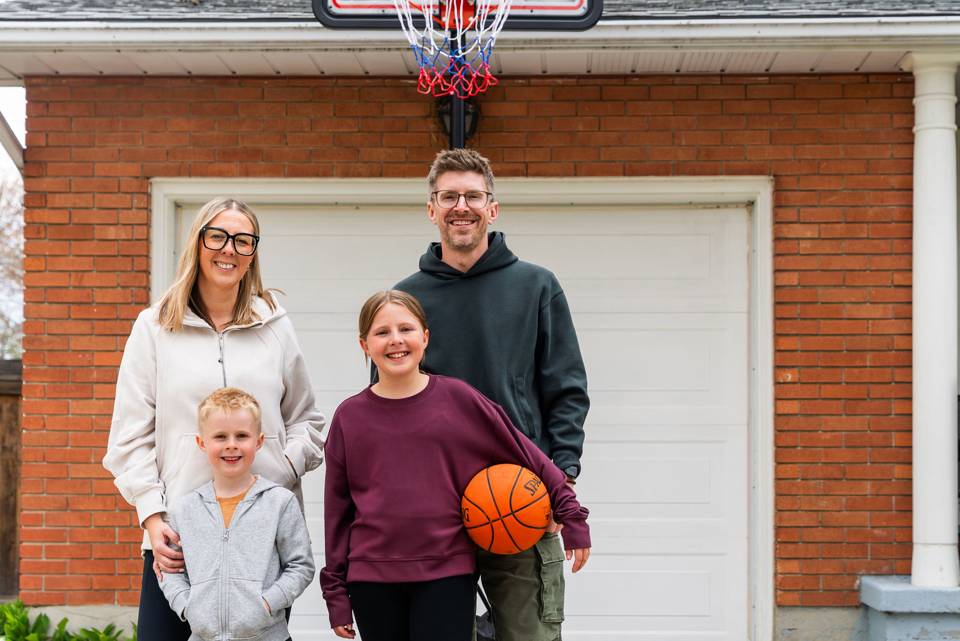 A family of four stands in front of a garage, with a basketball hoop above them. The girl holds a basketball while the boy stands beside her. The parents stand behind, smiling.