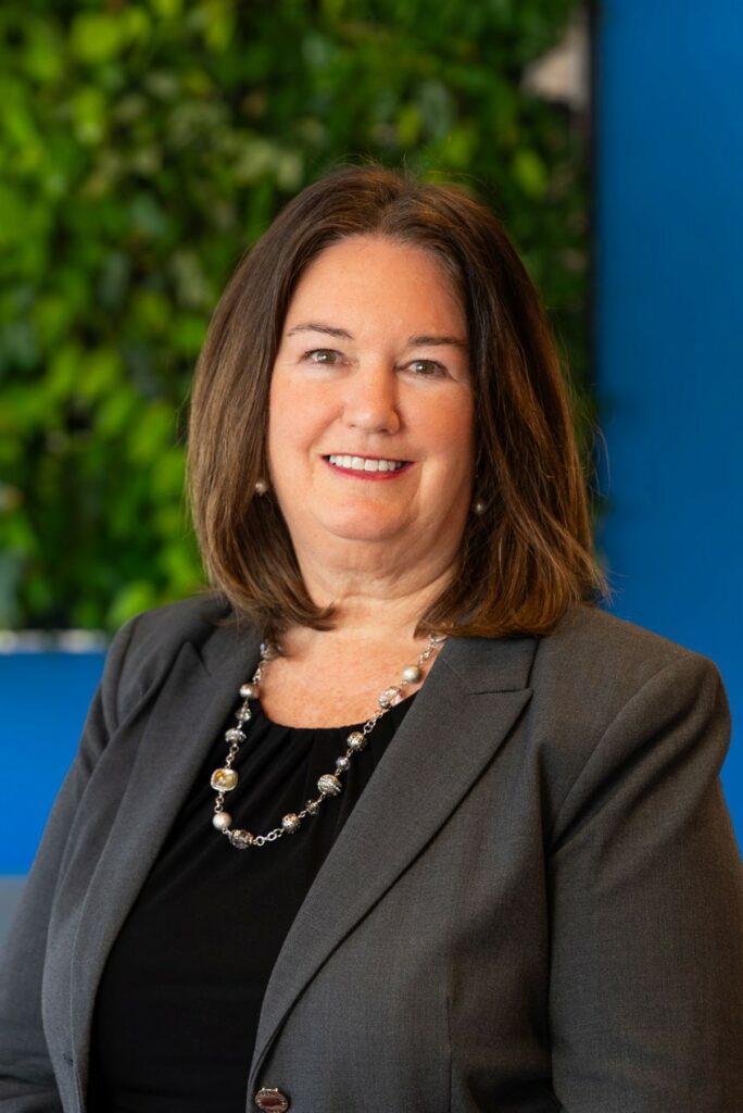 A woman with shoulder-length brown hair, wearing a gray blazer, black top, and beaded necklace, smiles in front of a green and blue background.