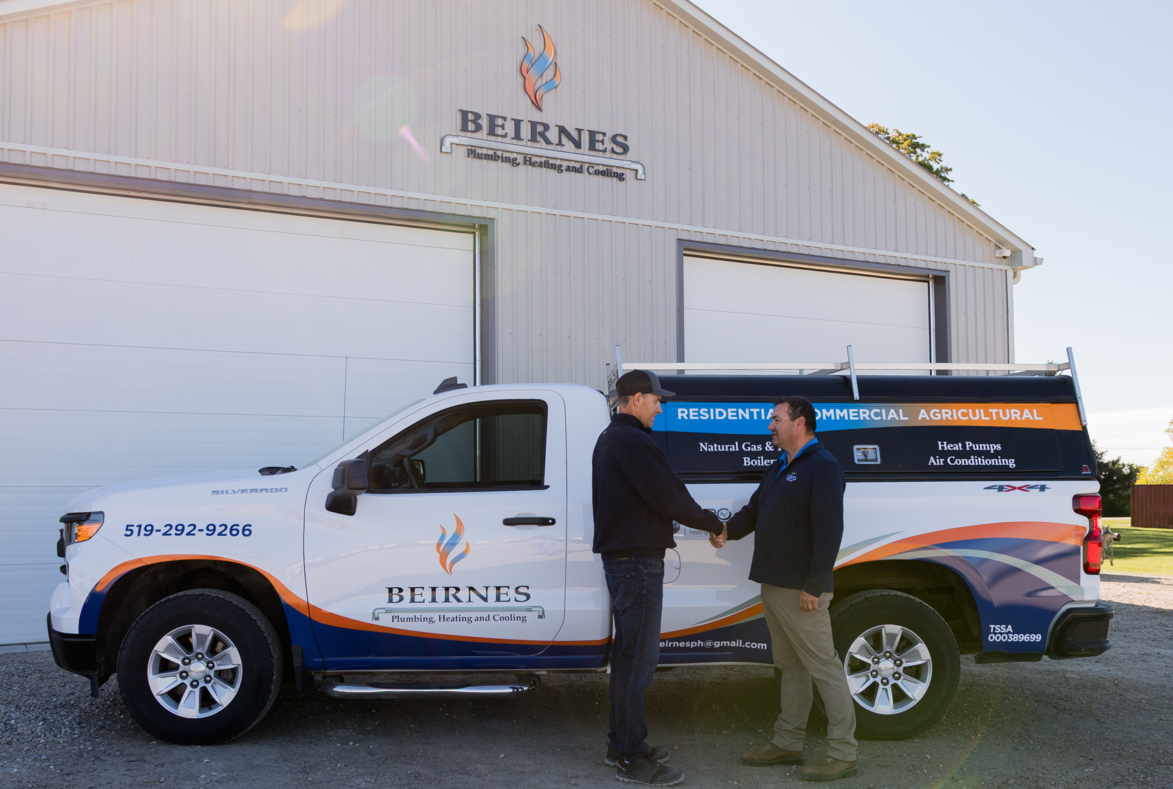 Two men in branded work shirts shake hands in front of a Beirnes Plumbing, Heating and Cooling service truck parked outside their shop.
